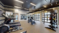 Located at 1340 Wilburn Blvd. in Orbetz, Ohio, this single-floor fitness center offers a thoughtfully designed workout environment that balances industrial aesthetics with modern functionality. The space is defined by its open-concept layout, providing generous room to move freely between equipment zones without feeling crowded.

Architecturally, the facility stands out with its exposed ductwork running along a dark, textured ceiling, complemented by sleek linear LED lighting that illuminates the floor evenly. Large glass-paned garage-style doors line one wall, flooding the interior with natural light and offering exterior views, while additional windows and glass entry doors further enhance the bright, airy atmosphere.

A bold, multi-toned striped accent wall serves as a striking visual focal point, adding a dynamic design element that sets this space apart from typical fitness facilities. Full-length mirrors along one wall visually expand the space and assist users during strength training exercises.

The equipment selection is comprehensive, including treadmills, stationary bikes, stair climbers, rowing machines, a multi-station cable machine, TRX suspension trainers, free weights, and medicine balls organized on dedicated racks. Durable rubberized flooring throughout provides comfort and safety. A separate connected room offers additional equipment and workout space, making this fitness center a versatile and well-appointed facility.
