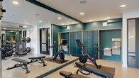 This fitness center offers a comprehensive exercise environment thoughtfully designed to support a wide range of workout routines. The space is divided into distinct functional zones, beginning with a cardio and strength area featuring treadmills, a multi-station cable machine, adjustable weight benches, and a full rack of dumbbells. An industrial-style exposed ductwork ceiling adds a modern edge to this zone, which also benefits from a large glass garage-style door that opens to an outdoor area, flooding the space with natural light and views of surrounding greenery. A wall-mounted flat-screen TV provides entertainment during workouts. The cycling studio area is outfitted with Peloton spin bikes positioned on floor mats, complemented by full-length mirrors that enhance the sense of space. Teal accent walls paired with warm wood-toned built-in storage cabinetry create a polished, contemporary aesthetic. A dedicated storage rack holds stability balls, resistance bands, and yoga mats, while a built-in water fountain station ensures convenient hydration. Recessed lighting throughout keeps the environment bright and welcoming, and durable light-toned flooring ties the entire space together with a clean, functional finish.