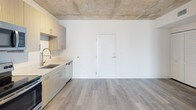 Located at 35 SW 6th Street in Miami's vibrant Brickell neighborhood, this single-floor unit blends industrial design with modern finishes to create a stylish and functional living space. The exposed concrete ceiling serves as a defining architectural feature, adding an urban loft character that contrasts beautifully with the crisp white walls and light wood-tone flooring found throughout.

The open living area flows seamlessly toward a floor-to-ceiling sliding glass door, framing impressive views of the Miami skyline and filling the space with abundant natural light. Track lighting mounted to the concrete ceiling provides additional illumination and highlights the unit's clean aesthetic.

The kitchen is well-appointed with light wood cabinetry, white quartz countertops, a stainless steel undermount sink with a flexible-head faucet, and a full suite of stainless steel appliances including a refrigerator, electric range, microwave, and dishwasher. A teal accent wall adds a bold pop of color to the living area, complementing the otherwise neutral palette.

The bedroom includes a closet with a built-in hanging rod, and connects conveniently to the bathroom, which features a soaking tub with large-format tile surround, a quartz vanity, and wood-look tile flooring. The layout is efficient and thoughtfully designed for comfortable city living in the heart of Miami.
