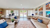 Located at 7263 Napa Valley Ln in Springdale, Arkansas, this single-floor community space offers a thoughtfully designed and inviting environment. The layout centers around a spacious living room that serves as the heart of the space, finished with rich hardwood-style flooring that runs throughout and crisp white crown molding that frames the neutral-toned walls.

Natural light fills the interior through multiple large windows fitted with blinds, as well as a set of glass double doors that open to an outdoor view, creating a seamless connection between the interior and the surrounding exterior. Recessed ceiling lights complement the natural light, ensuring the space remains bright and welcoming at all times.

The living area is comfortably arranged with seating, an area rug, and decorative wall art, contributing to a warm and functional atmosphere. A console table near the entry adds a practical yet stylish touch to the welcoming foyer area. A short hallway with additional wall art and an interior door leads to adjoining utility areas, adding to the overall functionality of the space.

The overall ambiance is clean, modern, and community-oriented, making this space well-suited as a gathering or common area. Its open feel, abundant natural light, and polished finishes combine to create an environment that is both practical and aesthetically appealing.