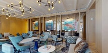 Located at 6–16 Livingston Avenue in New Brunswick, NJ, this single-floor lobby and amenity space serves as the welcoming centerpiece of Premier Residences, a luxury residential community. The entrance features floor-to-ceiling glass walls and bronze-framed double doors that flood the space with natural light while offering clear views of the surrounding streetscape.

The herringbone-patterned hardwood flooring runs throughout the entire space, lending a timeless, upscale character to the interior. Warm wood-paneled walls define a media lounge area, where built-in cabinetry and a large wall-mounted display screen create an inviting retreat for residents.

The main lounge area is generously proportioned and thoughtfully furnished with a mix of gray sofas, patterned accent chairs, and bold, colorful area rugs that add vibrancy and personality. A striking multi-bulb geometric chandelier anchors the space overhead, complementing the recessed lighting throughout. Vibrant large-scale artwork adorns the walls, contributing to the dynamic, curated atmosphere.

A circular upholstered seating arrangement near the glass entrance serves as an intimate conversation area, while a separate lounge section offers a more social setting with varied seating configurations. Decorative wall accents and sculptural lighting fixtures add further visual interest. The overall ambiance is modern, polished, and welcoming — reflecting a high standard of communal living.