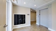 This building's common lobby area presents a clean, functional design characterized by neutral beige walls, warm-toned tile flooring, and a drop ceiling with recessed lighting that creates a bright and welcoming environment. A prominent built-in mailbox unit is mounted on the main wall, offering individual compartments for multiple units — a practical and organized solution for residents or occupants. Adjacent to the mailbox station, clear signage is posted on the wall, contributing to the orderly and informative nature of the space. To the right, an elevator with stainless steel doors provides convenient vertical access, accompanied by a wall-mounted control panel. A hallway extends toward the rear of the lobby, lined with additional recessed lighting and leading further into the building. White baseboards and door trim provide crisp contrast against the warm wall tones, lending the space a polished, well-maintained appearance. Overall, the lobby conveys a practical yet tidy aesthetic, offering essential amenities in a compact, efficiently arranged common area suitable for a multi-unit residential or commercial building.