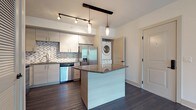 This well-appointed single-floor residence offers a thoughtfully designed open-concept layout centered around a modern kitchen and generous living area. The kitchen is a standout feature, showcasing light-toned cabinetry, granite countertops, a full suite of stainless steel appliances including a refrigerator with ice maker, electric range, over-the-range microwave, and dishwasher. A decorative mosaic tile backsplash adds visual texture, while a central island with a granite top provides additional prep space and casual seating. Pendant and track lighting illuminate the kitchen and entry area with a contemporary flair. A stacked washer and dryer unit is conveniently integrated into the space near the kitchen. Rich, dark hardwood-style flooring extends throughout the main living areas, lending warmth and continuity to the open floor plan. The living area is bright and airy, featuring a ceiling fan, large sliding glass doors that open to a balcony with lush outdoor views, and ample wall space. The bedroom offers plush carpeting, a window with natural light, and a ceiling light fixture. The bathroom is finished with neutral large-format tile, an open walk-in shower with matte black fixtures, and a soaking tub, creating a clean and modern aesthetic throughout.