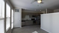 This single-story space showcases a contemporary open-concept layout that seamlessly connects the living and kitchen areas. The kitchen is a standout feature, equipped with a full suite of stainless steel appliances, including a French door refrigerator with ice and water dispenser, an electric range, a built-in microwave, and a dishwasher. Dark wood-toned cabinetry lines the walls, complemented by dark countertops that add a sleek, modern contrast to the neutral white walls. A single-basin sink is positioned along the main cabinet run, with a clean white tile backsplash completing the look. Recessed lighting illuminates the kitchen, while a ceiling fan with a light fixture serves the adjoining living area, enhancing air circulation and comfort. The flooring throughout is a wood-look plank style in a warm gray tone, providing a cohesive and low-maintenance surface. Natural light filters in through windows fitted with blinds, including a high transom window above an interior door, contributing to an airy feel. The overall ambiance is clean, modern, and functional.