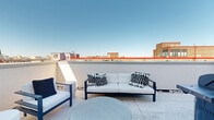 Located in the heart of Washington, DC, this rooftop terrace at 1224 M Street Northwest offers an exceptional outdoor living experience with sweeping panoramic views of the city skyline. The terrace is finished with large-format stone-look pavers that lend a clean, contemporary aesthetic throughout the open-air space.

The primary lounge area is generously proportioned, furnished with sleek navy-and-white outdoor seating — including a multi-seat sofa, accent chairs, and a sculptural round coffee table — arranged to take full advantage of the expansive city views. Decorative river rock borders accent the perimeter, adding a refined, low-maintenance landscaping detail.

A pergola-covered section near the sliding glass doors provides partial shade and a seamless indoor-outdoor transition, with floor-to-ceiling glazing that reflects the surrounding skyline. Modern wall-mounted light fixtures illuminate the space after dark, extending its usability into the evening hours.

A dedicated grilling zone, equipped with a full-sized gas grill and additional cooking equipment, makes the terrace well-suited for entertaining. Privacy walls and a slatted wood screen panel provide seclusion while preserving the open-air atmosphere. The clear sight lines across Washington's rooftops, including glimpses of notable landmarks, make this terrace a standout feature of the property.