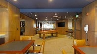 This single-floor recreational space is designed with entertainment and socializing in mind. At its centerpiece sits a full-size pool table with a rich orange felt surface, illuminated by two stylish pendant lights that hang from the dark ceiling above, drawing immediate visual focus to the area. The room is finished with warm wood paneling along the walls, lending a sophisticated and cohesive aesthetic throughout. Recessed lighting supplements the pendant fixtures, ensuring the space is well-lit across all areas. A wall-mounted flat-screen television is positioned prominently on one wall, complemented by a bar-height counter and stools nearby, creating a comfortable viewing and social area. Lounge seating and game tables are arranged around the perimeter, offering flexible options for both casual conversation and tabletop activities. Neutral-toned carpet tiles cover the floor, adding warmth underfoot while defining the space. A window at the far end of the room allows natural light to filter in, brightening the interior. The overall ambiance is modern yet inviting, balancing a club-like atmosphere with functional recreational amenities.