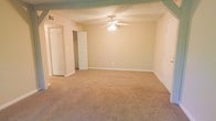 This single-floor residential space offers a clean and functional layout designed for comfortable everyday living. The main living area is generously sized, featuring wall-to-wall beige carpeting that adds warmth and comfort underfoot. A centrally mounted ceiling fan with a built-in light fixture provides both air circulation and ambient lighting, making the room feel practical and inviting. The textured ceiling adds a classic architectural detail throughout the space. Neutral beige walls create a versatile backdrop, allowing for a wide range of interior design styles and décor choices. The room benefits from multiple doorways, including access to what appears to be a hallway and additional rooms beyond. Storage is well-addressed, with louvered bifold closet doors visible along one wall, offering convenient and accessible organization options. White trim and baseboards run throughout, providing a crisp contrast to the warm-toned walls and flooring. The overall ambiance is bright, open, and move-in ready, with a straightforward and efficient use of space that suits a variety of living arrangements.