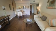 This single-floor residence is designed around an open-concept floor plan that integrates the living, dining, and kitchen areas into one cohesive, functional space. Rich, dark hardwood flooring runs throughout, grounding the interior with warmth and a sense of continuity.

The living area is anchored by a neutral-toned sofa accented with colorful throw pillows, paired with a round glass-top coffee table that adds a light, airy quality to the space. A tall lamp with a wood base and ceramic accent piece contributes to the warm, ambient lighting in the room. Framed artwork on the walls adds a decorative touch without overpowering the space.

Adjacent to the living area, the dining space features a white rectangular table surrounded by sleek white chairs, offering a clean and modern aesthetic. A pass-through opening with a mirrored backsplash connects the dining area to the kitchen, enhancing the sense of depth and openness while allowing natural interaction between the spaces.

The kitchen, visible through the pass-through, features light cabinetry and appears well-appointed. A hallway beyond leads to the remaining rooms, maintaining a logical and efficient flow throughout the residence.