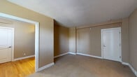 This single-floor residential unit offers a straightforward and functional layout designed for comfortable everyday living. The main living and bedroom areas are finished with neutral-toned wall-to-wall carpeting in a warm beige hue, complemented by tan and taupe wall colors that create a cohesive, understated aesthetic throughout. White baseboards provide a clean contrast against the warmer tones of the walls.

Natural light enters through windows fitted with horizontal blinds, offering both brightness and privacy control. A wall-mounted air conditioning unit is present in one of the carpeted rooms, ensuring year-round climate comfort.

The transition area features warm hardwood flooring, adding a classic and durable surface to the entry and connecting spaces. This area also benefits from a window with blinds, allowing natural light to filter in.

The bathroom is well-appointed with timeless finishes, including classic white subway tile surrounding a full bathtub and shower combination, penny-round white mosaic tile flooring, and a vanity topped with a rich granite-style countertop set in a dark wood cabinet. A window above the tub introduces natural light, while a large mirror and chrome fixtures complete the space.

A dollhouse view of the layout reveals a compact yet efficient floor plan, with the bathroom centrally accessible and a kitchen area visible in the upper portion of the unit, featuring cabinetry overhead. The overall ambiance is clean, neutral, and move-in ready.