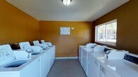 This shared laundry facility is a practical, well-organized space designed to serve the needs of its users efficiently. The room is lined on both sides with rows of white top-load washing machines and matching dryers, providing ample capacity for laundry needs. The walls are finished in a warm, bold golden-ochre tone, giving the space a distinct visual character, while the textured white ceiling adds a classic touch. Neutral gray flooring runs throughout, offering a clean and easy-to-maintain surface. A window on the right wall allows natural light to enter, brightening the interior and providing ventilation. Service information notices are clearly posted on the wall, ensuring users have easy access to instructions and contact details. Overhead ceiling-mounted lighting ensures the space remains well-lit at all times. The layout is thoughtfully arranged to allow users to move comfortably between machines on either side, making the process of doing laundry straightforward and convenient. Overall, this laundry room is a functional, no-frills communal facility that prioritizes practicality and ease of use.