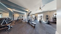 Located at 1640 W Elliot Rd in Gilbert, AZ, this single-floor fitness center offers a generously sized exercise space designed to accommodate a full spectrum of workout needs. The layout is open and thoughtfully organized, with distinct zones dedicated to cardio, strength training, and functional fitness.

The cardio section is well-equipped with multiple treadmills — including curved, self-powered models — alongside ellipticals, stationary bikes, and a rowing machine. Natural light streams in through numerous windows fitted with roller shades, creating a bright and energizing environment. Ceiling fans are positioned throughout to maintain comfortable airflow.

The strength training area features a multi-station cable machine, adjustable weight benches, and a full rack of dumbbells neatly arranged along a mirrored wall. The floor-to-ceiling mirrors visually expand the space while allowing users to monitor their form. An Octopus functional trainer system with resistance bands, weight plates, and accessories provides a versatile option for dynamic workouts.

The flooring consists of durable, multi-tone carpet tiles that define different workout zones, complemented by rubberized matting in high-use strength areas. The walls are finished in a neutral gray palette accented by slate-blue architectural details, lending a modern and professional aesthetic. A wall-mounted television adds an entertainment element to the space. The overall ambiance is clean, well-lit, and purpose-built for serious fitness use.