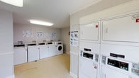 This shared laundry facility is designed with functionality and ease of use in mind. The single-floor space is outfitted with a row of front-loading washing machines along one wall and a bank of stacked dryers along the opposite side, providing ample capacity for multiple users. Each machine is clearly numbered for identification, and instructional posters — including guides for "Easy Washing" and "Simple Drying" — are posted prominently on the walls, ensuring residents can operate the equipment with confidence. The machines feature coin-operated or card-based payment controls, adding convenience to the laundry process. The room is finished with white walls accented by crown molding, lending a clean and polished appearance. Overhead fluorescent lighting ensures the space is consistently well-lit throughout the day. Light-toned flooring completes the bright, neutral aesthetic of the room. The overall layout is efficiently organized, maximizing the available floor space while maintaining clear walkways between equipment. The facility offers a practical and tidy environment that supports the everyday needs of its users.