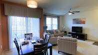 This single-floor residence offers an inviting open-concept layout where the dining and living areas flow naturally into one another, creating a cohesive and functional living environment. The dining area is anchored by a dark-stained wood table set with stylish place settings, complemented by matching chairs that add a touch of elegance to everyday meals. Large vertical-blind windows flood the space with abundant natural light, creating a bright and welcoming atmosphere throughout the day.

The living area extends seamlessly from the dining space, featuring plush seating including a comfortable sofa adorned with decorative accent pillows in warm tones, alongside patterned accent chairs that add visual interest and texture. A ceiling fan with a light fixture provides both comfort and illumination, while a flat-screen television rounds out the entertainment setup. Neutral-toned carpeting and warm wall accents in earthy hues contribute to the space's cohesive and cozy aesthetic.

Thoughtful design choices, including coordinated décor, layered lighting, and an accent wall in a rich terracotta tone, give the space a polished, well-curated feel. The abundance of windows ensures the interior remains airy and connected to the outdoors, making this a comfortable and visually appealing place to live.