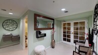 This single-floor wellness spa offers a thoughtfully designed environment for relaxation and therapeutic treatments. The entryway sets an inviting tone with soft sage-green walls, white crown molding, and classic small-format white tile flooring. A large ornate mirror, a modern floating shelf, and a tall decorative vase create an elegant first impression, while white French doors with glass panes provide a refined entrance into the space.

The massage room is a tranquil retreat, featuring warm neutral tones, crown molding, and recessed lighting that casts a soft, soothing glow. A professional treatment table takes center place, accompanied by a product shelf stocked with massage supplies and tropical-themed artwork that enhances the calming ambiance. A decorative framed mirror adds a finishing touch to the room.

A dedicated steam and sauna room is accessible through a frosted glass door, accompanied by a traditional wood-door sauna. Informational signage on the mint-green walls provides guidance for guests using the facilities.

The bathroom is clean and functional, featuring accessible design elements including a pedestal sink with grab bars and open under-sink clearance. White tile flooring ties the space cohesively together, ensuring practicality and ease of use throughout this well-appointed wellness facility.