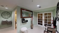 This single-floor wellness spa offers a thoughtfully designed environment for relaxation and therapeutic treatments. The entryway sets an inviting tone with soft sage-green walls, white crown molding, and classic small-format white tile flooring. A large ornate mirror, a modern floating shelf, and a tall decorative vase create an elegant first impression, while white French doors with glass panes provide a refined entrance into the space.

The massage room is a tranquil retreat, featuring warm neutral tones, crown molding, and recessed lighting that casts a soft, soothing glow. A professional treatment table takes center place, accompanied by a product shelf stocked with massage supplies and tropical-themed artwork that enhances the calming ambiance. A decorative framed mirror adds a finishing touch to the room.

A dedicated steam and sauna room is accessible through a frosted glass door, accompanied by a traditional wood-door sauna. Informational signage on the mint-green walls provides guidance for guests using the facilities.

The bathroom is clean and functional, featuring accessible design elements including a pedestal sink with grab bars and open under-sink clearance. White tile flooring ties the space cohesively together, ensuring practicality and ease of use throughout this well-appointed wellness facility.