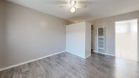 Located at 3734 W El Segundo Blvd in Hawthorne, California, this single-floor unit offers an efficient and functional layout encompassing a living room, kitchen, bathroom, and closet. The living room features durable wood-style laminate flooring in a warm gray tone, neutral painted walls with clean white baseboards, a ceiling fan with a light fixture, and a large window dressed with vertical blinds that allows natural light to fill the space. A warm-toned wood entry door adds a welcoming contrast to the room's neutral palette. A partial wall divides the living area, behind which a wall-mounted HVAC unit and a closet with open shelving provide added utility. The kitchen is thoughtfully appointed with white cabinetry — both upper and lower — granite-patterned countertops, a stainless steel sink, and a white gas range with four burners. White tile flooring grounds the kitchen space cleanly. The bathroom maintains a bright, all-white aesthetic with tile flooring, a combination tub and shower with a handheld showerhead, a white vanity with storage, a wall mirror, and a small window above the tub that introduces natural light. The overall design is clean, practical, and well-maintained throughout.