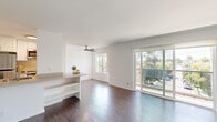 This well-appointed single-floor residence showcases a clean, modern aesthetic defined by crisp white walls, rich hardwood flooring, and an abundance of natural light throughout. The open-concept living and dining area serves as the heart of the space, anchored by a ceiling fan and framed by large sliding glass doors that lead to a private balcony overlooking a tree-lined street, filling the room with natural light and outdoor views.

A half-wall peninsula with open shelving elegantly separates the living area from the galley-style kitchen, which is outfitted with white shaker cabinetry, neutral quartz-style countertops, a stainless steel sink, and a full suite of stainless steel appliances including a refrigerator, range, and built-in microwave. The kitchen's efficient linear layout maximizes storage and counter space.

The bedroom is generously proportioned, featuring a ceiling fan with light fixture, a horizontal window providing natural light, and a full-width mirrored sliding closet that enhances the sense of space. Additional rooms maintain the same consistent palette of light wood flooring and white walls, contributing to a cohesive, airy ambiance. The overall design emphasizes openness, functionality, and a contemporary minimalist style throughout.