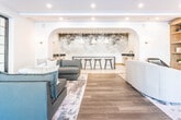 Located at 38 Old Short Hills Road in Livingston, New Jersey, this single-floor amenity space is thoughtfully designed to blend comfort and contemporary elegance. The open layout is anchored by warm hardwood flooring that flows seamlessly throughout, unifying the distinct zones within the space.

The main lounge area is generously proportioned, featuring a curated arrangement of plush seating, including rounded sofas and sleek accent chairs, centered on a textured area rug. Built-in wood shelving with display niches lines one wall, complemented by a textured, multicolored wallcovering that adds visual warmth and depth. Recessed lighting and wall sconces provide layered illumination throughout.

A dramatic arched opening frames the wet bar area, which is appointed with a dark island, backless bar stools, pendant lighting, and a striking abstract wall mural. This bar nook is flanked by cabinetry and a sink, making it well-suited for social gatherings.

A separate dining nook accommodates an intimate round table with dark leather chairs beneath a sculptural sputnik chandelier. Floor-to-ceiling grid windows and glass-panel doors throughout the space invite abundant natural light and provide views of the surrounding exterior. The overall ambiance is polished, modern, and welcoming.