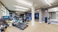 Located at 597 Westport Ave in Norwalk, CT, this fitness center offers a comprehensive and well-appointed workout facility across a single, generously sized floor. The space is divided into distinct functional zones, each designed to support a full range of exercise routines. The main cardio area features a row of treadmills, elliptical machines, and stationary bikes, all equipped with individual screens for an engaging workout experience. Large windows along one wall flood this section with natural light and provide views of the surrounding greenery, creating an inviting atmosphere. Structural columns with clean white detailing add a refined architectural touch throughout. Linear pendant lighting and recessed ceiling fixtures ensure bright, even illumination across the entire space. A dedicated free weights area includes a full dumbbell rack and benches, while a separate strength training zone — finished with durable speckled carpet flooring — houses weight plates, resistance equipment, and storage cabinetry. The accent walls, painted in a deep blue-gray tone, contrast stylishly with the neutral ceiling and trim, and wall-mounted mirrors throughout enhance the sense of depth and openness. Decorative artwork adds a polished, welcoming character to the space overall.