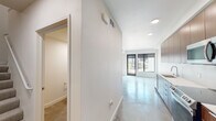 This contemporary two-floor space showcases a clean, modern aesthetic throughout. The open main level features polished concrete floors that reflect natural light, creating a bright and airy atmosphere. Large black-framed windows and a glass door at the front of the living area frame outdoor views while inviting an abundance of daylight into the space.

The galley-style kitchen is well-appointed with warm wood cabinetry extending to the ceiling, crisp white countertops, and a full suite of stainless steel appliances including a range, over-the-range microwave, and refrigerator. A white tile backsplash completes the clean, contemporary look.

Adjacent to the main living area, a bathroom is accessible from the hallway, offering a double-sink vanity with matching wood cabinetry and white countertops, a full wall mirror, and a combined tub and shower. The bedroom features polished concrete flooring, an exposed concrete ceiling detail adding an industrial character, and direct access to a walk-in closet with built-in shelving.

Throughout the space, white walls, recessed lighting, and consistent material choices create a cohesive and minimalist design. The two-floor layout provides a well-organized flow, making this a functional and stylish modern space.