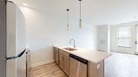 This thoughtfully designed single-floor space offers a clean, contemporary aesthetic carried consistently throughout. The open-concept kitchen serves as the heart of the unit, anchored by a spacious peninsula island topped with white quartz-style countertops and an undermount stainless steel sink with a brushed nickel faucet. Dual cylindrical pendant lights hang above the island, adding a modern design touch. The kitchen is fully equipped with stainless steel appliances, including a refrigerator, electric range, over-the-range microwave, and dishwasher, complemented by white shaker-style upper cabinets and warm-toned lower cabinetry. Light wood-look plank flooring extends seamlessly across the entire space, enhancing its bright and airy feel. The living area offers generous open space with white walls that maximize the sense of light and volume. A bedroom provides a calm, minimalist retreat with the same consistent flooring and neutral palette. The bathroom is well-appointed with a granite-topped vanity, warm-toned cabinetry, a large wall mirror, and a walk-in glass-enclosed shower with a built-in bench. A distinctive wood-finish entry door with a modern keypad lock adds a warm, contemporary accent at the entrance.