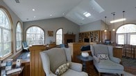 This single-floor clubhouse is thoughtfully designed to provide a warm and inviting communal gathering space. The main living area immediately draws attention with its soaring vaulted ceiling, accentuated by a pair of skylights that flood the room with natural light throughout the day. Large arched windows frame picturesque views of surrounding woodland, further enhancing the airy, open ambiance.

The space centers around a classic brick fireplace, serving as a natural focal point and adding a sense of warmth and character to the room. A built-in wooden bookcase with generous shelving lines one wall, offering both practical storage and a refined aesthetic. Rich wood paneling and brick-patterned flooring contribute to the space's traditional charm and architectural detail.

Comfortable wingback chairs and accent seating are arranged to encourage relaxed conversation, while thoughtful decorative touches add personality without overwhelming the space. Recessed lighting complements the natural light provided by the skylights, ensuring the area remains well-lit at all times. An adjacent area with cabinetry and pendant lighting suggests additional functional space for dining or catering. Overall, the clubhouse strikes a balance between casual comfort and classic elegance, making it an ideal venue for community gatherings and social events.