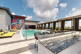 Located at 8448 Delmar Blvd in University City, Missouri, this rooftop amenity deck delivers a resort-caliber outdoor experience designed for relaxation and socializing. The centerpiece is a generously sized swimming pool with crystal-clear blue water, surrounded by expansive porcelain tile decking. Vibrant yellow chaise lounges line the poolside, offering ample space for sunbathing beneath open skies. A separate hot tub is tucked nearby, accented by a bold, large-scale artistic mural that commands attention along the building's exterior wall — its dynamic swirls of color and pattern providing a striking visual backdrop. A stylish poolside bar counter finished in veined marble-style stone offers a sophisticated spot for refreshments. Private cabana structures with slatted wood pergola roofing and elegant pendant chandeliers provide shaded retreats with lounge seating and personal television screens. A covered outdoor lounge area features comfortable sectional seating with open views of the surrounding landscape. Decorative topiaries and a standout gorilla topiary sculpture add a playful, artistic touch to the environment. The overall ambiance is modern and upscale, blending contemporary architecture with curated art and lush greenery to create a dynamic, resort-inspired outdoor destination.