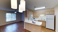 Located at 1 Dansey Ct in Durham, North Carolina, this single-floor residence offers a well-designed, open-concept layout that seamlessly connects the kitchen and living areas. The kitchen is a functional centerpiece of the home, featuring light wood cabinetry that provides ample storage, white appliances including a range, microwave, and dishwasher, and a peninsula with a clean white countertop that doubles as a casual dining or prep space. A double basin sink is positioned along the perimeter cabinetry, complemented by under-cabinet lighting that brightens the workspace. Overhead, pendant lighting with cylindrical shades adds a contemporary touch to the kitchen and adjoining dining area. Rich hardwood-style flooring runs throughout, lending warmth and cohesion to the open floor plan. The adjacent living area benefits from natural light streaming through a window, while a ceiling fan with a light fixture ensures year-round comfort. Neutral wall tones throughout the space create a clean, inviting atmosphere that is easy to personalize. The single-floor layout makes for effortless navigation, offering a practical and comfortable living environment in the heart of Durham.