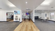 Located at 8330 N 19th Ave in Phoenix, Arizona, this fitness center at Montelano offers a generously sized, single-floor exercise room designed to support a full range of workout routines. The space is architecturally distinguished by soaring vaulted ceilings with a ceiling fan, large arched windows that flood the interior with natural light and provide views of the surrounding outdoor areas, and a clean white color palette that keeps the environment bright and energizing.

The flooring transitions between hardwood-style planks in the cycling and stretching zones and durable rubber matting in the strength and cardio areas, providing both function and visual contrast. A dedicated cardio section houses treadmills and an elliptical trainer, while a separate strength zone features a full dumbbell rack with a weight bench. Cycling enthusiasts will find multiple stationary spin bikes positioned in front of mounted flat-screen TVs for an engaging ride.

A standout functional training rig includes a heavy bag, kettlebells, medicine balls, resistance bands, and battle ropes, catering to high-intensity and cross-training workouts. An open mat area provides ample room for stretching and floor exercises. A water fountain with a bottle-filling station adds convenience. Decorative botanical wall murals lend the space a modern, polished aesthetic, rounding out this well-equipped community fitness facility.