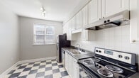 This single-floor space offers a cohesive and functional layout with consistent design details throughout. The living areas are defined by warm-toned hardwood floors, soft gray walls, crisp white baseboards, and elegant crown molding. Ceiling fans with integrated lighting fixtures are present in multiple rooms, providing both comfort and a classic aesthetic touch.

The main living room is generously proportioned, featuring a built-in alcove niche and a ceiling fan with a light kit. Natural light filters in through windows fitted with blinds, and the room opens naturally toward other areas of the unit. A second bedroom-sized room includes multiple windows on two walls, allowing ample natural light and views of surrounding greenery, along with its own ceiling fan. Another room features a closet and direct access to additional spaces.

The kitchen stands out with its striking black-and-white checkered tile flooring, white raised-panel cabinetry extending to the ceiling, dark countertops, a white subway tile backsplash, and a full suite of stainless steel appliances including a range, dishwasher, and refrigerator. A window above the sink area brings in additional natural light.

The bathroom is finished in an all-white palette, featuring white floor tiles, wainscoting, a bathtub with shower, and a window for ventilation and light.
