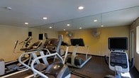 This fitness center is a dedicated, single-floor exercise space designed to meet a range of cardio and general workout needs. The room is outfitted with multiple treadmills and elliptical machines, each equipped with individual display screens for entertainment and performance tracking during workouts. A stationary bike is also available, rounding out the cardio equipment selection. Floor-to-ceiling mirrors line one full wall of the space, creating a sense of openness and allowing users to monitor their form while exercising. The mirrors also reflect the recessed overhead lighting, keeping the room bright and well-illuminated throughout. Two wall-mounted fans are positioned strategically to ensure adequate airflow and ventilation during intense exercise sessions. The flooring features durable, dark rubberized matting, which provides both safety and stability underfoot. The warm-toned walls contribute to an energizing yet focused atmosphere. The layout is practical and efficiently organized, allowing users to move between equipment with ease. Overall, this fitness center offers a clean, functional, and well-maintained environment suitable for daily cardio training and general exercise routines.