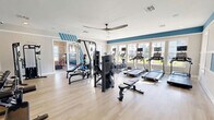 Located at 100 Vintage Alcoa Way in Alcoa, Tennessee, this single-floor fitness and activity center offers a thoughtfully designed space for health and recreation. The layout encompasses a generously sized exercise room and a separate activity room, making it a versatile amenity for residents.

The exercise room is bright and inviting, finished with light wood-style flooring and a crisp white and blue color scheme that runs along the upper walls. Abundant natural light streams in through a row of large windows, offering outdoor views and an energizing atmosphere. Ceiling fans help maintain airflow throughout the space. The room is well-equipped with multiple treadmills, elliptical machines, a multi-station cable machine, free weights with a dedicated rack, adjustable benches, and a full-wall mirror that enhances the sense of space.

The activity room features the same light flooring and a bold navy accent wall framing a large flat-screen television. Flanked by bright windows that bring in natural light and views of the surrounding brick exterior, this room is furnished with casual seating and is well-suited for recreation and leisure. Crown molding and a clean white wainscoting detail add a polished, residential feel throughout both spaces, contributing to an overall atmosphere that is functional yet welcoming.
