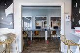 The Business Center is a thoughtfully designed single-floor space that balances functionality with contemporary aesthetics. The main workspace area features a long granite countertop workstation equipped with desktop computers, accommodating multiple users simultaneously. Elegant gold-framed chairs provide comfortable seating, while large pass-through windows overlook an adjacent lounge area, creating a sense of openness and visual connectivity throughout the space.

Rich hardwood-style flooring runs throughout, complementing the crisp white walls adorned with bold black-and-white abstract artwork. The center benefits from generous natural light, particularly in the secondary lounge and meeting area, where sliding glass doors open directly onto an outdoor deck with views of surrounding greenery.

A well-appointed kitchenette serves the space, featuring white cabinetry with brushed hardware, a double sink, granite countertops, a full-size stainless steel refrigerator, microwave, and toaster oven. Two round gold-framed mirrors above the counter add a decorative touch. The overall ambiance is polished and modern, with consistent gold metallic accents tying together the furnishings and décor throughout the entire space.