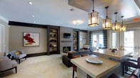 This single-floor space showcases a refined, open-concept design that seamlessly connects the living and dining areas. The living room serves as a focal point, anchored by a sleek fireplace framed in herringbone-patterned tile and flanked by dark-framed built-in shelving units, which provide both display space and functional storage. A wall-mounted television is integrated above the fireplace, contributing to the clean, contemporary aesthetic.

The flooring features a striking herringbone-laid tile or stone pattern in warm neutral tones, adding texture and visual interest underfoot. Overhead, a tray ceiling detail defines the living space, complemented by recessed lighting and a decorative flush-mount fixture. Pendant lantern-style light fixtures with amber glass panels hang above the dining or kitchen counter area, lending a warm, layered glow to the space.

The kitchen island or bar counter is finished in a light, smooth surface, paired with upholstered seating in rich brown tones. Large French doors near the seating area invite natural light and offer views to the exterior. The overall ambiance is sophisticated and contemporary, combining dark accents with neutral tones for a polished, cohesive interior.