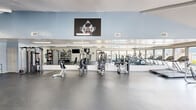 Located at 2700 Whitney Ave in Harvey, Louisiana, this fitness center offers a single-floor exercise room designed to support a comprehensive workout experience. The space features a clean, modern aesthetic with light blue and white walls, durable dark flooring, and vaulted ceilings in sections that lend an open, airy feel to the interior. Large windows along one side of the room invite abundant natural light and provide outdoor views of the surrounding grounds, creating a bright and motivating environment. A full-length mirror spans one wall, visually expanding the space and allowing users to monitor their form during exercise. The room is well-equipped with a varied selection of strength training machines, including cable and resistance units, as well as a pull-up and dip station. Cardio equipment such as upright and recumbent stationary bikes, ellipticals, and treadmills are positioned to take advantage of the natural light from the windows. A dedicated dumbbell rack provides additional free weight options. Multiple wall-mounted flat-screen televisions are positioned throughout the space for entertainment during workouts, and a motivational display is prominently featured on one wall. A water station is available for convenience. The facility also includes direct exterior access through a glass door.