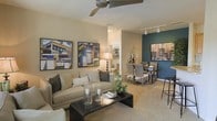 This single-floor residence offers a thoughtfully designed open-concept layout that seamlessly connects the living and dining areas into one cohesive, inviting space. The living area is anchored by a plush, neutral-toned sofa and a sleek dark coffee table adorned with decorative candle holders and greenery, creating a warm and welcoming atmosphere. A ceiling fan with a brushed metal finish overhead provides both function and style, while an abundance of soft, neutral wall tones give the space an airy, comfortable feel. Bold, colorful artwork lines the walls, adding a contemporary and artistic character to the room. Adjacent to the living area, the dining space is defined by a striking teal accent wall that serves as a dramatic focal point. A compact dining table paired with woven high-back chairs provides an intimate setting for meals, while track lighting above ensures the area is well-lit and practical. Soft carpeting flows throughout, contributing to the overall warmth of the interior. The open sightlines between the living and dining zones enhance the sense of spaciousness, making this single-floor residence as functional as it is visually appealing — ideal for both everyday living and entertaining.