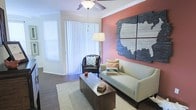 This single-floor living space combines contemporary design elements with warm, inviting touches to create a comfortable and visually engaging environment. The living room serves as the focal point, showcasing a bold coral-toned accent wall adorned with a striking large-scale mixed-media wall art piece depicting a map of the United States rendered in wood and slate-textured panels — an eye-catching architectural detail that anchors the room.

Dark hardwood-style flooring runs throughout the space, adding depth and contrast against the lighter furnishings and crisp white crown molding that frames the room. A ceiling fan with integrated lighting provides both style and function overhead. Natural light floods the interior through large sliding glass doors fitted with vertical blinds, which also offer access to an outdoor area. An additional window further enhances the brightness of the space.

The living area is thoughtfully arranged with a light-toned sofa, leather accent chair, a rectangular wood coffee table, and a woven area rug, creating a cohesive and functional seating arrangement. A floor lamp adds supplemental ambient lighting, contributing to the overall warm and welcoming atmosphere of the space.