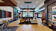 This contemporary amenity center makes a striking first impression with its soaring double-height entrance foyer, floor-to-ceiling glass walls, and warm hardwood flooring that flows throughout the space. The grand lobby anchors the facility with an open, airy layout bathed in natural light, featuring a dramatic pendant chandelier, a welcoming seating arrangement of neutral sofas and amber accent chairs, and expansive glazed walls that frame views of the surrounding landscape.

Moving deeper into the space, multiple distinct lounge areas offer varied atmospheres for socializing or relaxing. Rich, jewel-toned furnishings — including bold red and orange sofas — are set against deep plum accent walls, reclaimed wood paneling, and large black-framed windows that invite natural light and outdoor views. Wood-paneled ceiling insets and globe pendant lighting add warmth and a refined, layered character to each zone.

A dedicated co-working area provides a productive environment with workstation desks, ergonomic seating, and generous window views. Adjacent, a fully equipped fitness center is visible through sleek floor-to-ceiling glass panels, showcasing cardio and strength training equipment alongside textured reclaimed wood accent walls. Throughout the entire facility, the design seamlessly blends industrial and contemporary elements, creating an inviting, well-appointed communal environment.