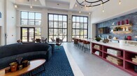 Located at 555 Dresher Rd in Horsham, PA, this single-floor community clubhouse showcases a contemporary design with soaring ceilings and an open, airy layout throughout. The centerpiece of the space is a dramatic main lounge bathed in natural light from floor-to-ceiling black-framed grid windows that frame views of the surrounding community. A striking circular chandelier with Edison-style pendant bulbs anchors the room, casting warm ambient light over the seating area, which features a curved navy velvet sofa, swivel lounge chairs, and a patterned blue area rug set atop large-format tile flooring. A bold magenta kitchen island with a crisp white countertop serves as a social hub, with bar seating along one side. Adjacent to it, a well-appointed beverage station offers coffee, tea, and refreshments, accented by a geometric blue tile backsplash and natural wood cabinetry. A glass-enclosed co-working room with black-framed sliding doors provides a quiet workspace visible from the main lounge. Additional seating nooks include a fireplace lounge with textured tile surround and a serene alcove with tall wingback chairs beneath curated wall art. A secondary lounge with plush armchairs and carpet extends toward exterior views, completing this thoughtfully designed communal retreat.