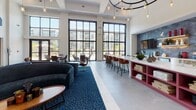 Located at 555 Dresher Rd in Horsham, PA, this single-floor community clubhouse showcases a contemporary design with soaring ceilings and an open, airy layout throughout. The centerpiece of the space is a dramatic main lounge bathed in natural light from floor-to-ceiling black-framed grid windows that frame views of the surrounding community. A striking circular chandelier with Edison-style pendant bulbs anchors the room, casting warm ambient light over the seating area, which features a curved navy velvet sofa, swivel lounge chairs, and a patterned blue area rug set atop large-format tile flooring. A bold magenta kitchen island with a crisp white countertop serves as a social hub, with bar seating along one side. Adjacent to it, a well-appointed beverage station offers coffee, tea, and refreshments, accented by a geometric blue tile backsplash and natural wood cabinetry. A glass-enclosed co-working room with black-framed sliding doors provides a quiet workspace visible from the main lounge. Additional seating nooks include a fireplace lounge with textured tile surround and a serene alcove with tall wingback chairs beneath curated wall art. A secondary lounge with plush armchairs and carpet extends toward exterior views, completing this thoughtfully designed communal retreat.