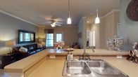 This single-floor residence offers a thoughtfully designed open-concept layout that effortlessly connects the kitchen, dining, and living areas into one cohesive, welcoming space. The kitchen serves as the heart of the home, featuring a double-basin stainless steel sink, a wraparound countertop that transitions into a breakfast bar, and stylish pendant lighting that adds a warm, modern touch overhead. The open flow of the layout creates a natural gathering space, ideal for both everyday living and entertaining. The living area is comfortably appointed, anchored by a ceiling fan that provides both function and charm, and is filled with warm, ambient light from a window dressed with blinds and curtains. Crown molding runs throughout the space, lending a refined, polished character to the interior. Neutral tones on the walls and countertops contribute to the space's calm and inviting atmosphere. A hallway visible in the background suggests additional private rooms beyond the main living area, reinforcing the practical and well-organized nature of the floor plan. Overall, the space strikes a balance between comfort and elegance, making it a functional and attractive place to call home.