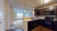 Located at 9220 Worthington Rd in Westerville, Ohio, this single-floor unit offers a well-organized layout designed for comfortable everyday living. The open-concept kitchen flows seamlessly into the living area, creating a connected and functional space. The kitchen is outfitted with rich, dark espresso cabinetry, light-toned countertops, a stainless steel sink set into a peninsula, and black appliances including an electric range and over-the-range microwave. Three warm amber pendant lights hang above the kitchen peninsula, providing both task lighting and a welcoming ambiance. Hardwood flooring anchors the kitchen and hallway areas, while plush carpeting extends through the living room and bedrooms, adding warmth underfoot. The living room benefits from natural light through a large window fitted with horizontal blinds. The bedroom is a generously sized, carpet-floored room with its own window offering exterior views. The bathroom features tile flooring, a dark-cabineted vanity with a light countertop, and direct access to a carpeted walk-in closet complete with built-in shelving and hanging rods. Throughout the unit, neutral warm tones on the walls create a cohesive and inviting atmosphere.