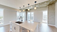 Located in Nashville's vibrant Midtown neighborhood, this one-bedroom, one-bathroom residence blends contemporary design with industrial-inspired architectural details. The open-concept living, dining, and kitchen area serves as the heart of the home, showcasing light hardwood floors, exposed concrete columns and ceiling accents, and expansive floor-to-ceiling windows that flood the space with natural light and frame sweeping city skyline views. The kitchen is well-appointed with white shaker cabinetry, quartz countertops, stainless steel appliances, a subway tile backsplash, and a large center island with a matte black faucet, illuminated by globe pendant lights overhead. Sliding glass doors lead to a private balcony, extending the living space outdoors. The bedroom offers plush carpeting, a ceiling fan, a large window with city views, and two closets for ample storage. The bathroom features a white quartz vanity, undermount sink, matte black fixtures, and a framed mirror. A hallway with built-in shelving and bench adds functional storage near the entry. In-unit laundry completes this well-designed urban residence.