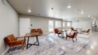 Located at 925 E Omaha St in Rapid City, South Dakota, this thoughtfully designed single-floor space offers a seamless open-concept layout that integrates a living room, dining area, and kitchen into one cohesive environment. The polished concrete floors run throughout, lending a clean, industrial-modern aesthetic that pairs well with the warm neutral wall tones and crisp white baseboards. Recessed lighting illuminates the space evenly, enhancing its bright and welcoming atmosphere. The living area is anchored by a striking feature wall in slate gray, complete with a sleek linear fireplace topped by a rustic wood mantel and wall-mounted display, alongside open wooden shelving that adds warmth and character. The dining and lounge areas are generously proportioned, offering multiple seating arrangements ideal for gathering, and benefit from natural light streaming through several windows and a glass-paneled exterior door that opens to an outdoor area. The kitchen is outfitted with modern gray flat-front cabinetry, white countertops, stainless steel appliances including a range with a wall-mounted hood, and a full-size refrigerator, providing ample functionality. The overall ambiance is contemporary, clean, and versatile — well-suited for communal or multi-purpose use.