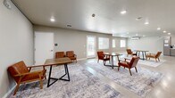 Located at 925 E Omaha St in Rapid City, South Dakota, this thoughtfully designed single-floor space offers a seamless open-concept layout that integrates a living room, dining area, and kitchen into one cohesive environment. The polished concrete floors run throughout, lending a clean, industrial-modern aesthetic that pairs well with the warm neutral wall tones and crisp white baseboards. Recessed lighting illuminates the space evenly, enhancing its bright and welcoming atmosphere. The living area is anchored by a striking feature wall in slate gray, complete with a sleek linear fireplace topped by a rustic wood mantel and wall-mounted display, alongside open wooden shelving that adds warmth and character. The dining and lounge areas are generously proportioned, offering multiple seating arrangements ideal for gathering, and benefit from natural light streaming through several windows and a glass-paneled exterior door that opens to an outdoor area. The kitchen is outfitted with modern gray flat-front cabinetry, white countertops, stainless steel appliances including a range with a wall-mounted hood, and a full-size refrigerator, providing ample functionality. The overall ambiance is contemporary, clean, and versatile — well-suited for communal or multi-purpose use.