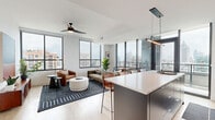 This contemporary 3-bedroom, 2-bathroom residence offers a thoughtfully designed open-concept layout spread across a single floor. The combined kitchen and living area serves as the heart of the home, anchored by large floor-to-ceiling windows that flood the space with natural light and frame impressive city skyline views. A ceiling fan adds a functional accent overhead, while light hardwood flooring runs throughout, lending warmth to the modern aesthetic.

The kitchen is outfitted with sleek, dark matte cabinetry extending to ceiling height, a white quartz countertop, white subway tile backsplash, and a full suite of stainless steel appliances. A spacious island with seating provides an ideal spot for casual dining or entertaining. The kitchen opens directly to the living area, which benefits from panoramic city views and access to a private balcony.

The three bedrooms are well-proportioned, with the primary bedroom offering dedicated closet space. Each room features the same light hardwood flooring and large windows with city views. The two bathrooms are cleanly finished — one features a soaking tub with subway tile surround, while the other offers a glass-enclosed walk-in shower. Both include dark vanity cabinetry and white quartz countertops. Two hallways connect the bedrooms and common areas, completing this well-organized urban residence.