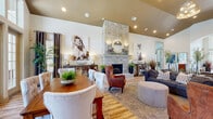This single-floor community clubhouse is designed with an upscale, welcoming aesthetic that blends comfort and functionality throughout. The centerpiece of the space is a sweeping main lounge boasting soaring vaulted ceilings, warm wood-look flooring, and an impressive floor-to-ceiling stacked stone fireplace that anchors the room with dramatic visual impact. A gold chandelier and recessed lighting illuminate the open-concept layout, which is furnished with tufted wingback chairs, a plush sofa, ottomans, and area rugs, creating an inviting gathering environment. Large multi-pane windows and glass French doors flood the interior with abundant natural light and provide views to the outdoors. A dedicated dining area with a farmhouse-style table and upholstered seating flows seamlessly into the lounge. A high-top bar table with industrial metal stools offers a casual seating option near the windows. The kitchen area features a granite-topped island with bar seating, open shelving, and stainless steel appliances. A business center is equipped with multiple computer workstations beneath stylish pendant lighting, complemented by a cozy seating lounge. The fitness center is well-appointed with cardio machines, free weights, spin bikes, wall-mounted televisions, and a large ceiling fan, rounding out this thoughtfully designed communal facility.