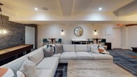 Located at 44 Hill St in Morristown, NJ, this thoughtfully designed community room offers a refined yet welcoming atmosphere ideal for social gatherings and recreation. The open-concept layout is anchored by a generous lounge area furnished with a large sectional sofa, accent armchairs in slate blue, and a rustic wood coffee table, all set atop a geometric-patterned carpet that introduces pops of teal and orange throughout the space. A wall-mounted flat-screen TV serves as a central focal point, complemented by recessed lighting and crown molding that lend the room a polished, upscale feel. Adjacent to the lounge, a game area features a full-sized pool table beneath a stylish pendant light, alongside a textured stone accent wall that adds warmth and visual depth. A wet bar with under-counter refrigeration, bar-height seating, and pendant lighting provides an inviting spot for entertaining. Small clerestory windows allow natural light to filter in, brightening the neutral palette of warm whites and soft grays. The restroom is a standout feature, showcasing reclaimed wood plank walls, white subway tile wainscoting, geometric patterned floor tile, a floating sink, and matte black fixtures — a striking blend of rustic and contemporary design that elevates the overall character of the space.