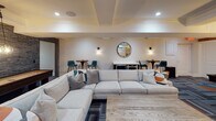 Located at 44 Hill St in Morristown, NJ, this thoughtfully designed community room offers a refined yet welcoming atmosphere ideal for social gatherings and recreation. The open-concept layout is anchored by a generous lounge area furnished with a large sectional sofa, accent armchairs in slate blue, and a rustic wood coffee table, all set atop a geometric-patterned carpet that introduces pops of teal and orange throughout the space. A wall-mounted flat-screen TV serves as a central focal point, complemented by recessed lighting and crown molding that lend the room a polished, upscale feel. Adjacent to the lounge, a game area features a full-sized pool table beneath a stylish pendant light, alongside a textured stone accent wall that adds warmth and visual depth. A wet bar with under-counter refrigeration, bar-height seating, and pendant lighting provides an inviting spot for entertaining. Small clerestory windows allow natural light to filter in, brightening the neutral palette of warm whites and soft grays. The restroom is a standout feature, showcasing reclaimed wood plank walls, white subway tile wainscoting, geometric patterned floor tile, a floating sink, and matte black fixtures — a striking blend of rustic and contemporary design that elevates the overall character of the space.