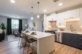 This thoughtfully designed single-floor residence offers a seamless blend of modern aesthetics and functional living. At the heart of the home is an open-concept kitchen and living area, where light wood flooring flows throughout and connects the spaces effortlessly. The kitchen is a standout, featuring a striking two-tone cabinetry design with crisp white upper cabinets and deep navy lower cabinets, complemented by a white subway tile backsplash with under-cabinet lighting. A large waterfall-edge island with a quartz marble-look countertop provides generous prep space and casual seating, while stainless steel appliances and contemporary pendant lighting complete the sophisticated look. The living area is bright and inviting, with natural light streaming in through large windows dressed in dark curtains, creating a warm yet stylish atmosphere. Two well-proportioned bedrooms offer comfortable retreats; each features plush carpet flooring, natural light from generously sized windows, and a calm, neutral color palette. The bathroom is finished with the same wood-look flooring, a dark navy vanity with a white countertop, a large mirror, and an elongated walk-through layout. Recessed lighting throughout the unit ensures a bright, cohesive ambiance across all areas.